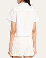 The backside view of a dark-haired model wearing a white, short-sleeved, button-up shirt with a collar and white shorts in front of a white background.