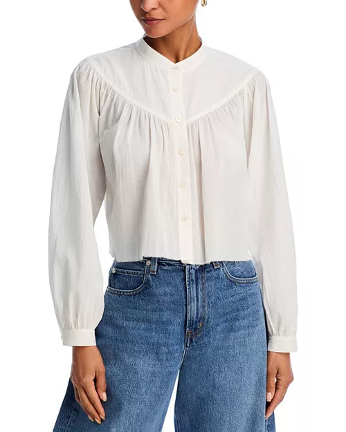 A torso shot of a model wearing a white long-sleeve blouse with a high collar and button-down front, a yoke design at the shoulders and loose-fitting sleeves that gather at the cuffs. The person is also wearing blue jeans and is standing in front of a white background.