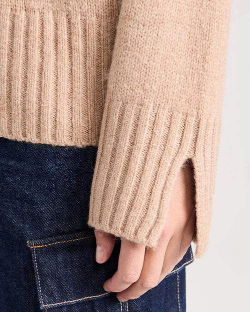 A close-up of a models' hand wearing a beige knitted sweater with ribbed cuffs and a wrist slit. The model is also wearing dark blue denim jeans with a side pocket.