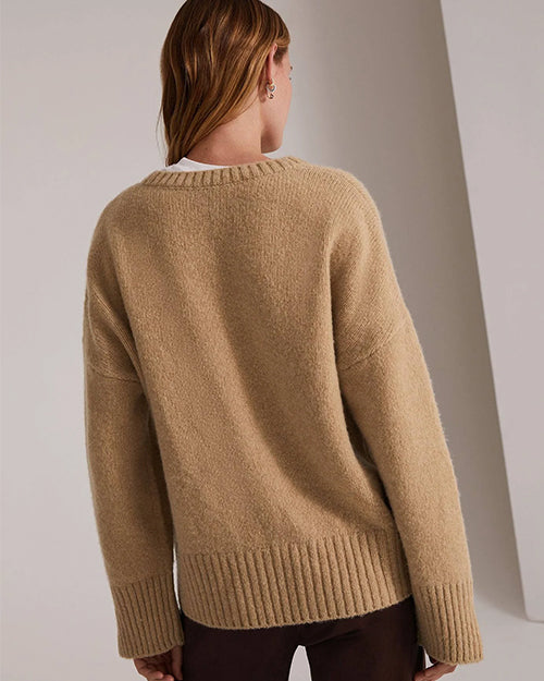 A knee-up backside view of a blonde model with her hair swept to the side wearing a beige V-neck sweater over a white t-shirt and dark brown trousers. The background is grey.