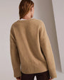 A knee-up backside view of a blonde model with her hair swept to the side wearing a beige V-neck sweater over a white t-shirt and dark brown trousers. The background is grey.