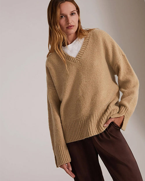 A knee-up view of a blonde model with her hand in her pocket wearing a beige V-neck sweater over a white t-shirt and dark brown trousers. The background is grey.