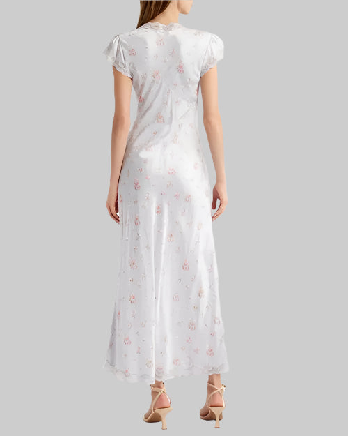 The backside view of a brunette model with her hair swept to the side wearing a long light-blue dress featuring short sleeves with laced edges and a floral pattern with small pink flowers. The model paired the dress with beige high-heels and is standing in front of a gray background.