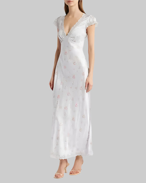 A body shot of a model wearing a long light-blue dress featuring short sleeves, a laced V-neckline, and a floral pattern with small pink flowers. The person is also wearing light colored heels and standing in front of a grey background.
