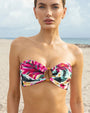 An upper body shot of a model standing on a sandy beach, wearing a strapless bandeau style bikini top with a colorful floral brushstroke pattern and an O-ring at the center.
