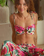A sitting model wearing a thin strapped bandeau style bikini top with a colorful floral brushstroke pattern, an O-ring design in the center, and bows tied at the straps.  She is wearing matching drawstring pants, and sitting in front of a potted plant and mirror in a living room setting.