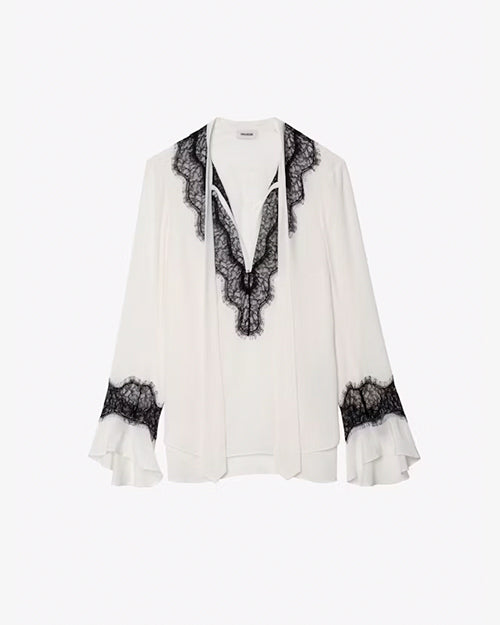 A full view of a white blouse with black lace detailing with scalloped edges around the V-neck neckline and around the lower end of the long sleeves with flared cuffs. The background is plain white.