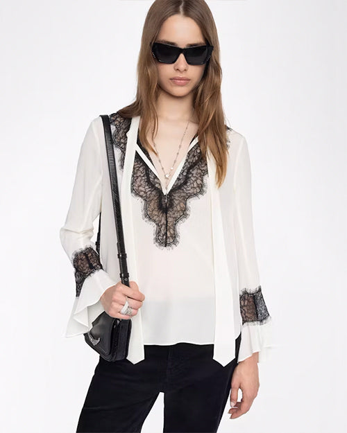 A knee-up view of a brunette model wearing a white blouse with black lace detailing around the V-neck neckline and around the lower end of the long sleeves with flared cuffs. The sleeves have flared cuffs. The model is also wearing black pants, black sunglasses, and carrying a black shoulder bag. The background is plain white.
