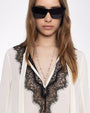 A close-up view of a brunette model wearing a white blouse with black lace detailing with scalloped edges around the V-neck neckline, and long white strings hanging down from the neck. The model is also wearing black sunglasses and a beaded silver necklace.