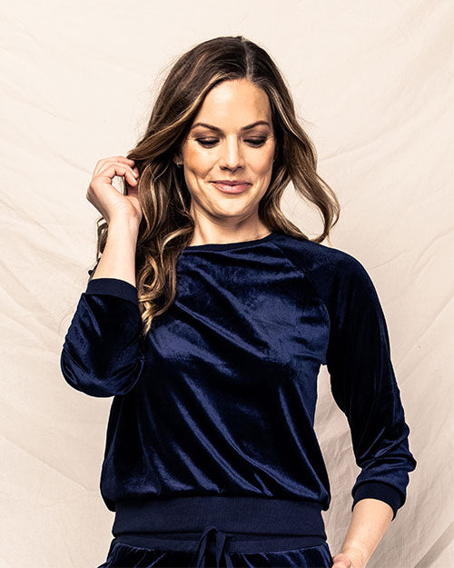 A waist- image of a model with one hand in her pocket and one hand up to her ear, wearing a navy blue long-sleeve pajama set against a white fabric background.