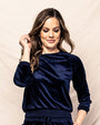 A waist- image of a model with one hand in her pocket and one hand up to her ear, wearing a navy blue long-sleeve pajama set against a white fabric background.