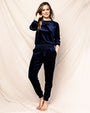 A full body image of a barefoot model with one hand in her pocket and one hand up to her ear, wearing a navy blue long-sleeve pajama set against a white fabric background.