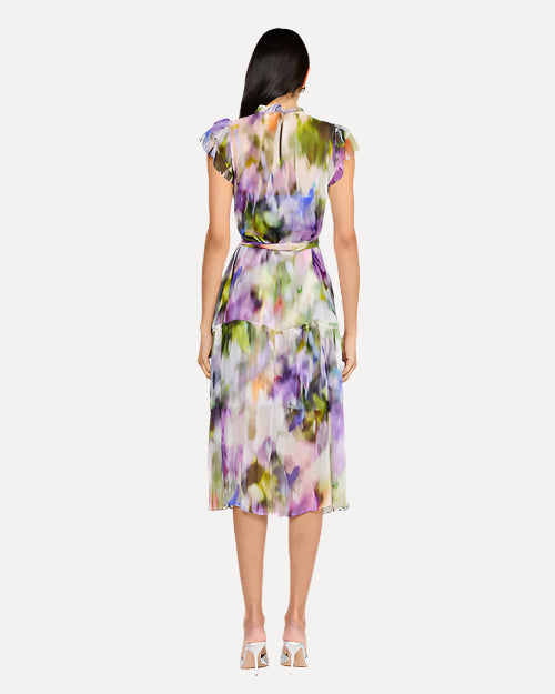 A backside view of a dark-haired model wearing a sleeveless, knee-length dress with a colorful abstract pattern, a high neckline with ruffled straps, and is cinched at the waist with a matching fabric belt. The model is also wearing white heels and is in front of a white background.