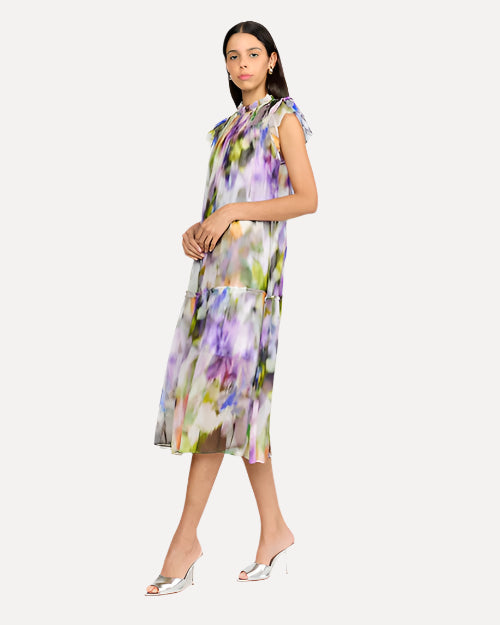 A side view of a dark-haired model wearing a sleeveless, knee-length dress with a colorful abstract pattern, a high neckline with ruffled straps and is loosely fitting. The model is also wearing white open-toe heels and is standing against a plain background.