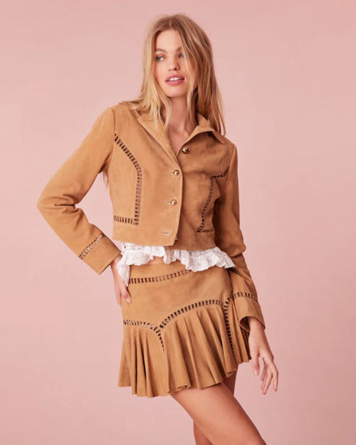 A knee-up view of a blonde model with her hand on her hip, dressed in a matching tan suede outfit consisting of a long-sleeved button-down jacket and a matching pleated skirt, adorned with decorative stitching featuring empty space rectangles. The model is wearing a white blouse under and is standing in front of a pink background.
