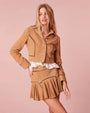A knee-up view of a blonde model with her hand on her hip, dressed in a matching tan suede outfit consisting of a long-sleeved button-down jacket and a matching pleated skirt, adorned with decorative stitching featuring empty space rectangles. The model is wearing a white blouse under and is standing in front of a pink background.