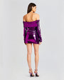 A full body backside view of a dark-haired model wearing a purple sparkly off-the-shoulder dress with long sleeves. The model is accessorizing with round gold earrings and pink high heels. The background is plain white.