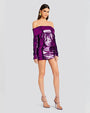 An angled full body of a dark-haired model wearing a purple sparkly off-the-shoulder dress with long sleeves. The model is accessorizing with round gold earrings and pink high heels. The background is plain white.
