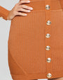 A close-up hip-level view of a model with her hand on her hip, wearing an orange long sleeve dress with gold buttons down the front and a ribbed material.