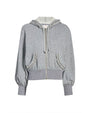 A gray zip-up hoodie with a drawstring hood and long sleeves. The hoodie features ruffled trim detailing along the edge of the hood and around the pocket openings. It has a ribbed waistband and cuffs, creating a slightly cropped and fitted silhouette.