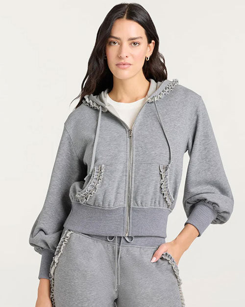 A gray zip-up hoodie with a drawstring hood and long sleeves. The hoodie features ruffled trim details along the hood edge, pocket openings, and waistband. It is paired with matching gray sweatpants that also have ruffled accents near the pockets. A white T-shirt is visible underneath the hoodie.