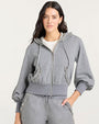 A gray zip-up hoodie with a drawstring hood and long sleeves. The hoodie features ruffled trim details along the hood edge, pocket openings, and waistband. It is paired with matching gray sweatpants that also have ruffled accents near the pockets. A white T-shirt is visible underneath the hoodie.