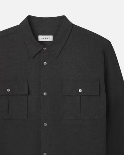 A close-up view of a dark long-sleeve silver button-up shirt with two chest pockets, and a rectangular white tag with the brand name "FRAME" on it. The background is plain white.