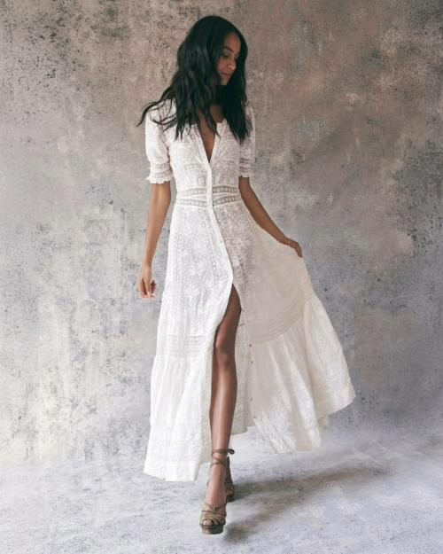 A full body view of a dark-haired model holding one end of a long, white lace dress with elbow-length ruffled, slightly puffed sleeves and striped lace detailing, also appearing on the waistline. The dress also has a V-neckline, and a side slit. The model is also wearing a pair of beige open-toed heels and standing against a grey splotchy background.