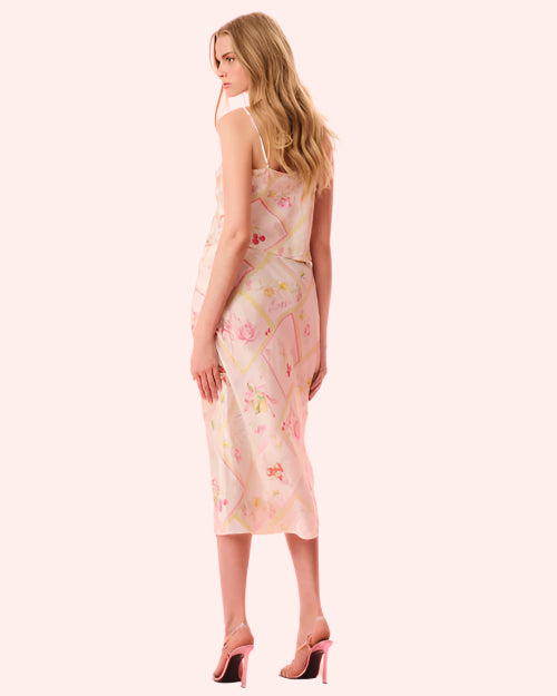 The back view of a woman wearing a matching white tank top and skirt, featuring vibrant fruit and floral patterns inside of colorfully outlined diamond shaped designs. The model is also wearing pink high heels against a plain background.