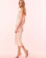 A sideview of a model wearing a matching white tank top and shin length skirt featuring vibrant fruit and floral patterns inside of colorfully outlined diamond shaped designs. The model is also wearing pink high heels against a plain background.