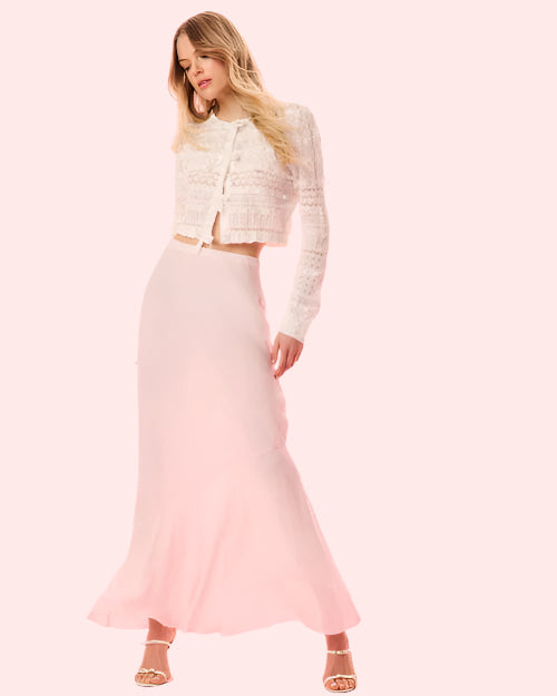 A blonde model standing next to a white background, dressed in a white lace cardigan with intricate detailing and a light pink skirt paired with white open-toed heels.