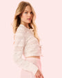 A blonde model in front of a white background showcasing a white lace cardigan with intricate detail all over it, paired with light pink bottoms.