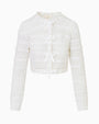 A white cropped cardigan featuring delicate lace detailing and lace bows down the front for closure, placed in front of a white background.