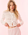 A blonde model poses in front of a light background in a white lace cardigan paired with white bottoms. The cardigan has intricate lace detailing and multiple bows down the front for closure.