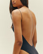 A close waist-up backside view of a brunette model wearing a black one-piece swimsuit with a completely open back and thin straps, standing in front of a white background.