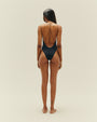 A full body backside view of a brunette model wearing a black one-piece swimsuit with a completely open back and thin straps, standing in front of a white background.