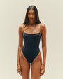 A knee-up view of a brunette model wearing a black one-piece swimsuit with a seam at the upper waist and thin straps, standing in front of a white background.