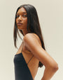A close waist-up backside view of a brunette model wearing a black one-piece swimsuit with a completely open back and thin straps, standing in front of a white background.