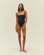 A full body view of a brunette model wearing a black one-piece swimsuit with a seam at the upper waist and thin straps, standing in front of a white background.