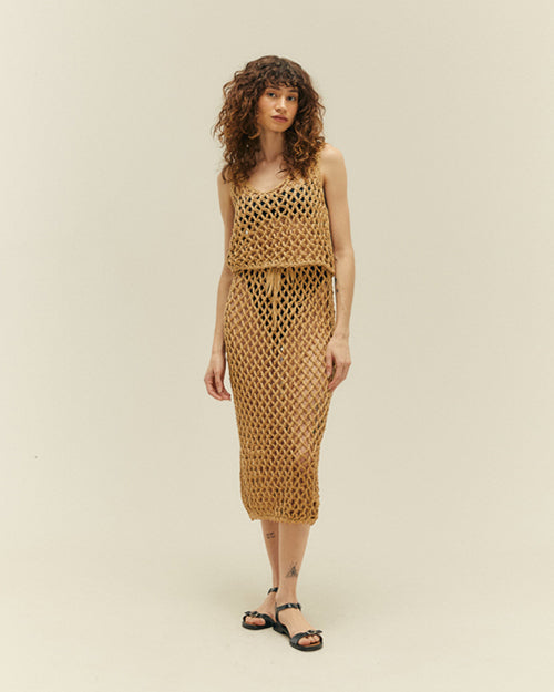 A full body view of a brunette model wearing a matching beige, diamond rope-knit tank top and a matching skirt with a bow tied at the top. The model is wearing a black bikini underneath with black open-toed sandals and is standing in front of a white background.