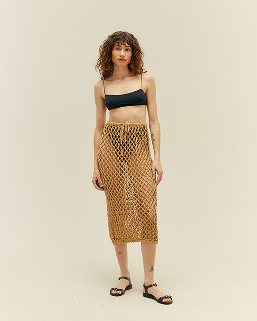 A full body view of a brunette model wearing a black bikini top with thin straps, and black bikini bottoms with a diamond rope-knit skirt over it, featuring a bow tie in the top. The model is also wearing black open-toed heels and is standing in front of a white background.