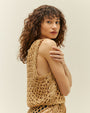 An angled sideview of a brunette model with her arms wrapped around her torso wearing a matching beige, diamond rope-knit tank top and matching bottoms. The model is wearing black bikini bottoms and is standing in front of a white background.