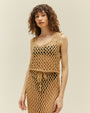 An angled view of a brunette model wearing a matching beige, diamond rope-knit tank top and a matching skirt. The model is wearing a black bikini underneath and is standing in front of a white background.