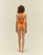 A full body backside view of a brunette model with her hair pushed to the side, wearing a bikini showcasing high-waisted orange bottoms and X-strapped back with a bow. The background is plain white.