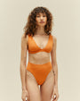 A knee-up view of a brunette model wearing a bikini showcasing a high-waisted orange bottoms and a V-neck top. The background is plain white.