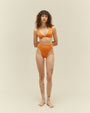 A full body view of a brunette model with leg tattoos and her arms behind her back, wearing a bikini showcasing a high-waisted orange bottoms and a V-neck top. The background is plain white.