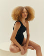 A blonde model sitting with one leg folded, wearing a black one-piece swimsuit with a slanted neckline and one strap with a bunched design at the top. The background is plain white.