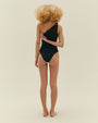 A full body backside view of a blonde model with her hand touching her back, wearing a black one-piece swimsuit with a slanted neckline and one strap with a bunched design at the top. The background is plain white.