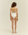 A full body backside view of a barefoot brunette model wearing a white one-piece swimsuit with an ovalis open back design. The background is plain white.
