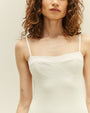 A close waist-up angle of a brunette model wearing a white one-piece swimsuit with a seam at the upper chest and thin straps, standing in front of a white background.
